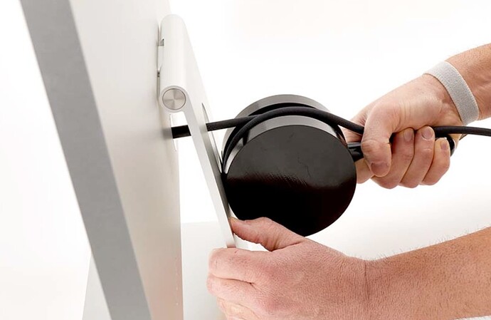 Side view of a Studio Display with the power cord wrapped around a large, black cylinder and one hand holds the Display's stand while the other tightly grips the power cable.